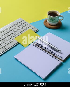 Blue and yellow tabletop with keyboard, notepad, pen, coffee and blank yellow paper. Stock Photo
