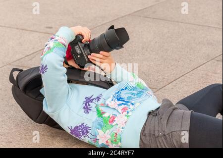 A very young female photographer lays on her back while using a large DSLR camera to get the perfect angle.