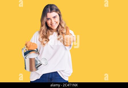 Young caucasian woman wearing gloves holding watering can smiling happy and positive, thumb up doing excellent and approval sign Stock Photo