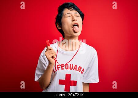 Young beautiful asian lifeguard girl wearing t-shirt with red cross using whistle sticking tongue out happy with funny expression. Emotion concept. Stock Photo