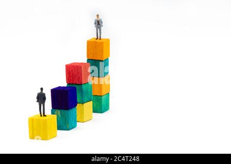 Miniature figurine posed as businessman sitting alone on a wooden cube looking up to leader on colourful stacks as business chart with focus on backgr Stock Photo