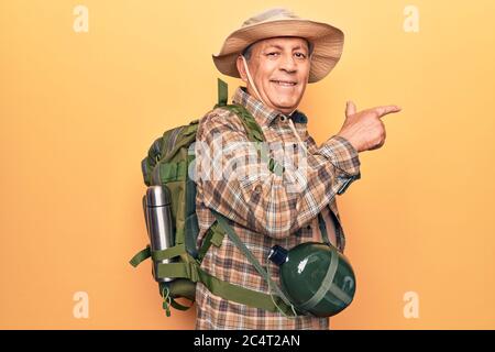 Senior man with grey hair wearing hiker backpack and water canteen smiling cheerful pointing with hand and finger up to the side Stock Photo