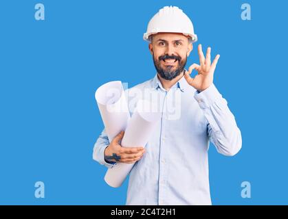 Young handsome man wearing hardhat holding paper blueprints doing ok sign with fingers, smiling friendly gesturing excellent symbol Stock Photo