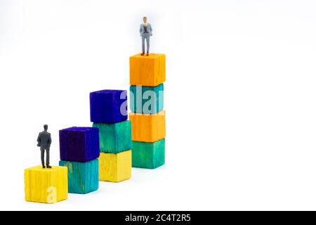 Miniature figurine posed as businessman sitting alone on a wooden cube looking up to leader on colourful stacks as business chart with focus on foregr Stock Photo