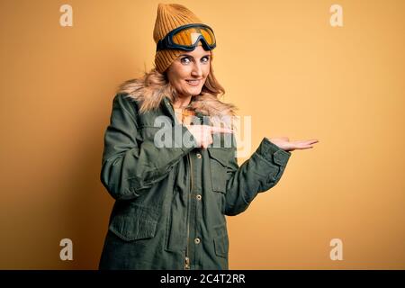 Middle age beautiful blonde skier woman wearing snow sportwear and ski goggles amazed and smiling to the camera while presenting with hand and pointin Stock Photo