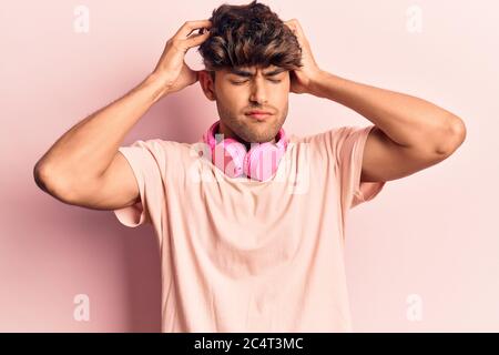 Young hispanic man listening to music using headphones with hand on head, headache because stress. suffering migraine. Stock Photo