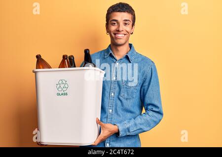 Young african amercian man holding recycling wastebasket with glass looking positive and happy standing and smiling with a confident smile showing tee Stock Photo