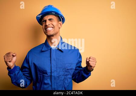 Young handsome african american worker man wearing blue uniform and security helmet very happy and excited doing winner gesture with arms raised, smil Stock Photo