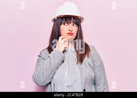 Young plus size woman wearing architect hardhat thinking concentrated about doubt with finger on chin and looking up wondering Stock Photo