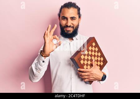 Young arab man holding chess doing ok sign with fingers, smiling friendly gesturing excellent symbol Stock Photo