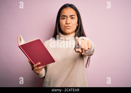 Young asian smart woman holding book diary standing over isolated pink background pointing with finger to the camera and to you, hand sign, positive a Stock Photo