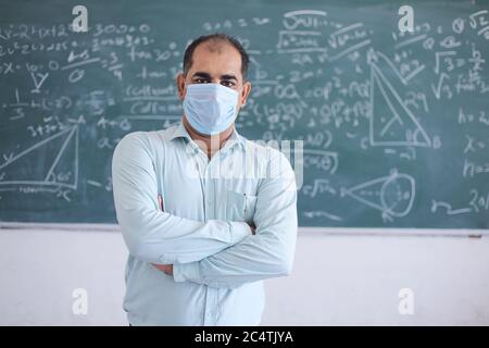 Portrait of male teacher wearing mask standing against blackboard teaching mathematics in classroom, school reopen after lockdown due to covid-19 pand Stock Photo