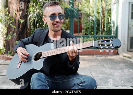 Smiling handsome man in sunglasses sitting on pavement and playing guitar Stock Photo