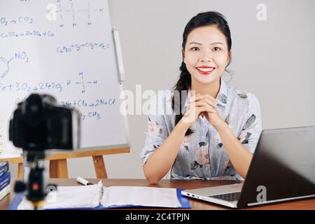 Portrait of positive young chemistry teacher sitting at desk when recording lesson for her students Stock Photo