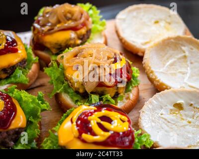 Tasty homemade juicy burger. Caramelized onion on mustard and ketchup on beef, cheese and fresh green lettuce on wood cutting board, process of making Stock Photo