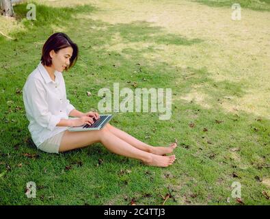 Beautiful Asian women wearing white shirt and shorts working with laptop computer sitting on green grass in park. Happy female outdoor working with te Stock Photo
