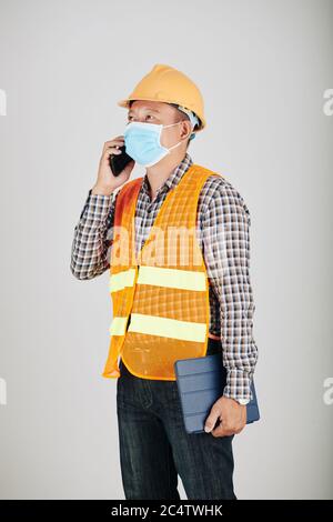 Portrait of Asian contractor in medical mask talking on phone with head engineer Stock Photo