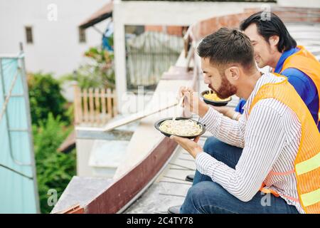 Builders sitting on second floor of building under constrution and eating noodles for lunch Stock Photo