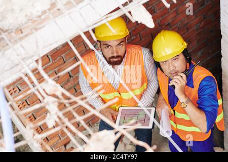 Builders working in building under construction, one holding tablet computer with blueprint, his colleague using with walkie-talkie Stock Photo