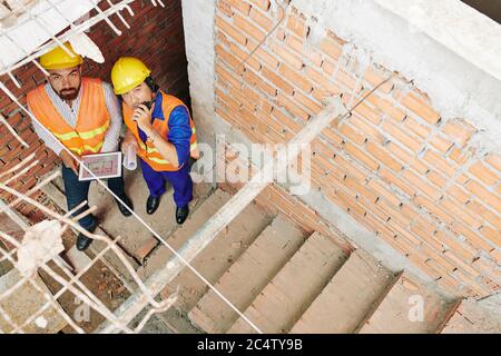 Contractors in handhats standing at staircase flight and using walkie-talking to communicate with workers on the roof Stock Photo