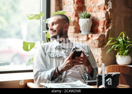 Smiling handsome young man in denim jacket sitting at table and texting message while waiting for friend in cafe Stock Photo