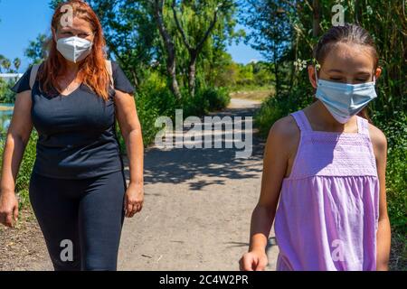 Middle-aged red-haired woman with her young daughter walking on a path surrounded by nature with masks to protect themselves from the coronavirus. Hea Stock Photo