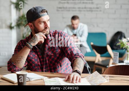 Pensive young bearded man sitting at table in cafe and looking away while thinking about new trip Stock Photo