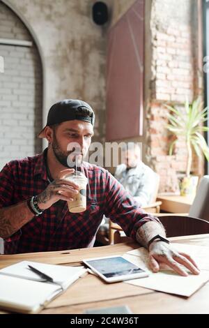 Serious focused young bearded man with tattoos sitting at table in cafe and drinking cocktail while planning trip using map Stock Photo
