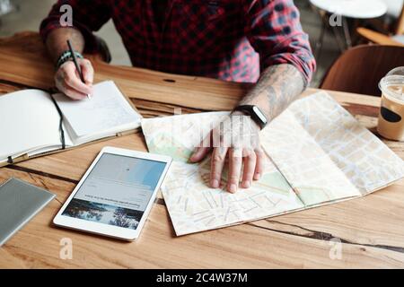 Close-up of unrecognizable tattooed man sitting at table in cafe and checking hotel location on map while planning trip Stock Photo