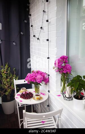 Bouquet of peony flowers, two cups of tea and fresh cherries on white table. Modern balcony interior. Stock Photo
