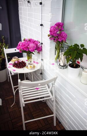 Bouquet of peony flowers, two cups of tea and fresh cherries on white table. Modern balcony interior. Stock Photo