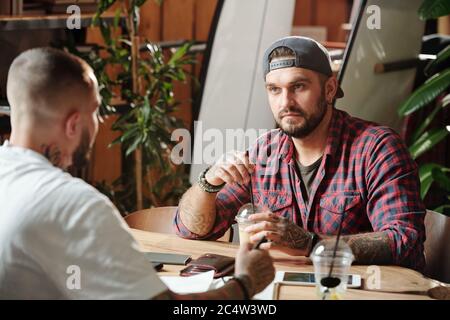 Brutal young men sitting at table in cozy cafe and drinking refreshing beverages while talking about life Stock Photo