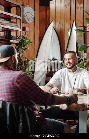 Handsome young man with tattoos sitting at table in cozy cafe and enjoying meeting with friend Stock Photo