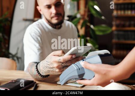Close-up of man with tattoos paying with smartphone after dinner in modern cafe Stock Photo