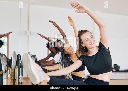 Smiling young men and women stretching legs on barre to warm up before training in dance class Stock Photo