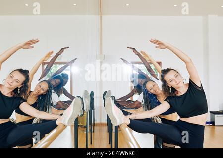 Pretty smiling young dancers putting feet on barre when doing stretching exercise for legs Stock Photo