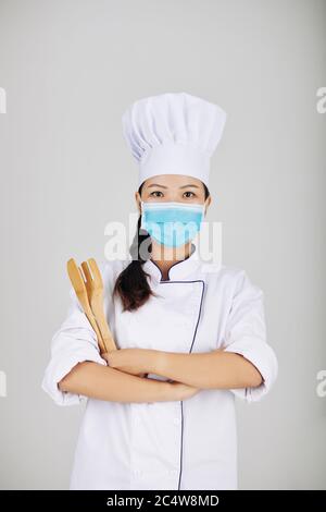 Portrait of young female chef in medical mask holding wooden fork and spatula when posing against grey backgrounf Stock Photo