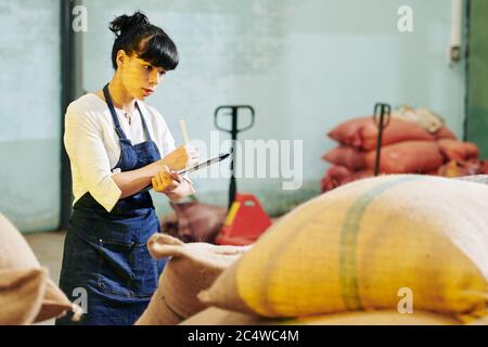 Young female warehouse worker checking sacks and taking notes in document on clipboard Stock Photo