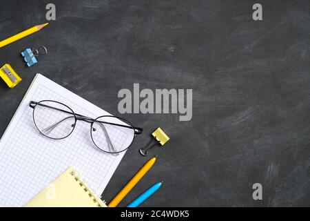 Back to school concept. Blank paper notebook, glasses and school supplies on black board background. Flat lay, top view, copy space. Stock Photo