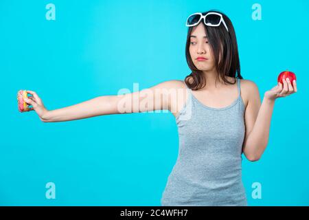 Attractive Asian young woman holding and choosing between disk of donuts or red apple fruit on isolated blue color background, weight loss and avoid j Stock Photo