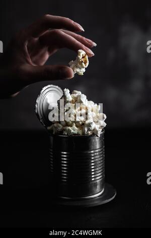 Popcorn in a steel tin on dark background, hand with a piece of popcorn, moody photo Stock Photo