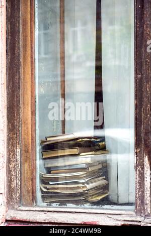 Old dilapidated hardcover books are stacked on a window sill near the window, close-up view from the outside through the glass. Stock Photo