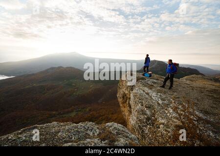 Two hikers with backpacks stand on the top of the mountain and looking at beautiful yellow autumn landscape sunset over clouds. Climbers relax on Stock Photo
