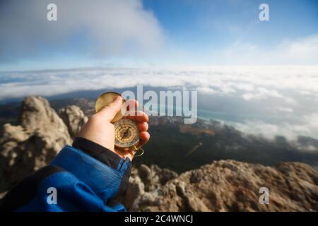 Point of view photo of explorer man searching direction with golden compass in his hand above clouds with autumn mountains and sea background Stock Photo