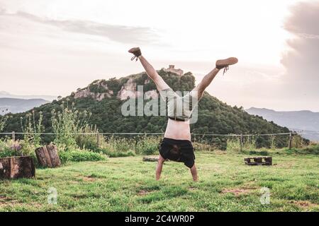Adventurous young man doing a handstand in the middle of nature. There is a mountain with a building on top. Stock Photo