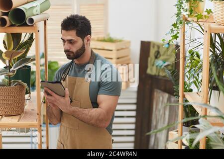 Waist up portrait of bearded man wearing apron using digital tablet while counting stock in flower store, copy space Stock Photo