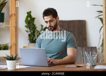Portrait of handsome bearded man using laptop while sitting at desk in home interior decorated with houseplants, copy space Stock Photo