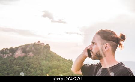 Handsome young man in profile talks on his cell phone. There is a mountain with a building on top. Stock Photo
