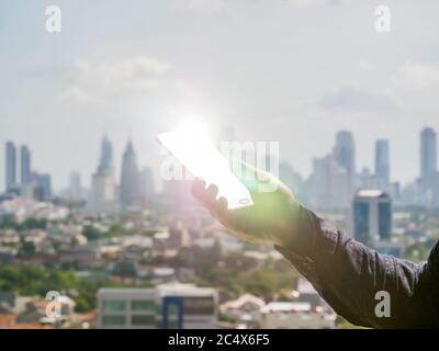A man holds a phone in his hand that shines a ray of sunshine against the background of the city of Jakarta. Indonesia. Stock Photo