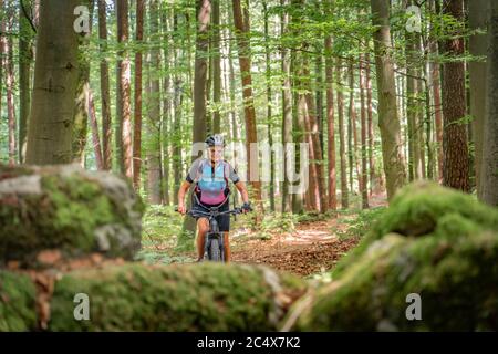 pretty senior woman underway on her electric mountain bike on a rocky forest trail in Franconian Switzerland, Bavaria, Germany Stock Photo